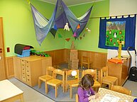 Eine Spielecke des Kindergartens. Braune Stühle bilden einen Halbkreis, auf dem Boden davor sitzt ein kleines Mädchen.