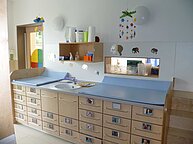 Das Bade und Wickelzimmer des Kindergartens. Man sieht ein Spülbecken und braune Kommoden mit blauen Tischplatten. 