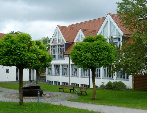 Das Bild zeigt das neue, moderne Gebäude des Kindergarten Merchings mit vielen Bäumen und vielen Fenstern.